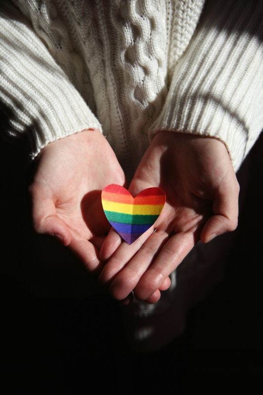 Photo of two hands holding out a paper heart with the rainbow pride flag